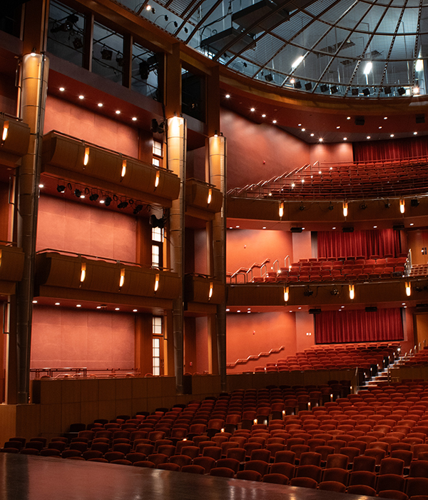 Interior of a modern theater auditorium with rows of red seats, multiple balconies, and warm stage lighting.